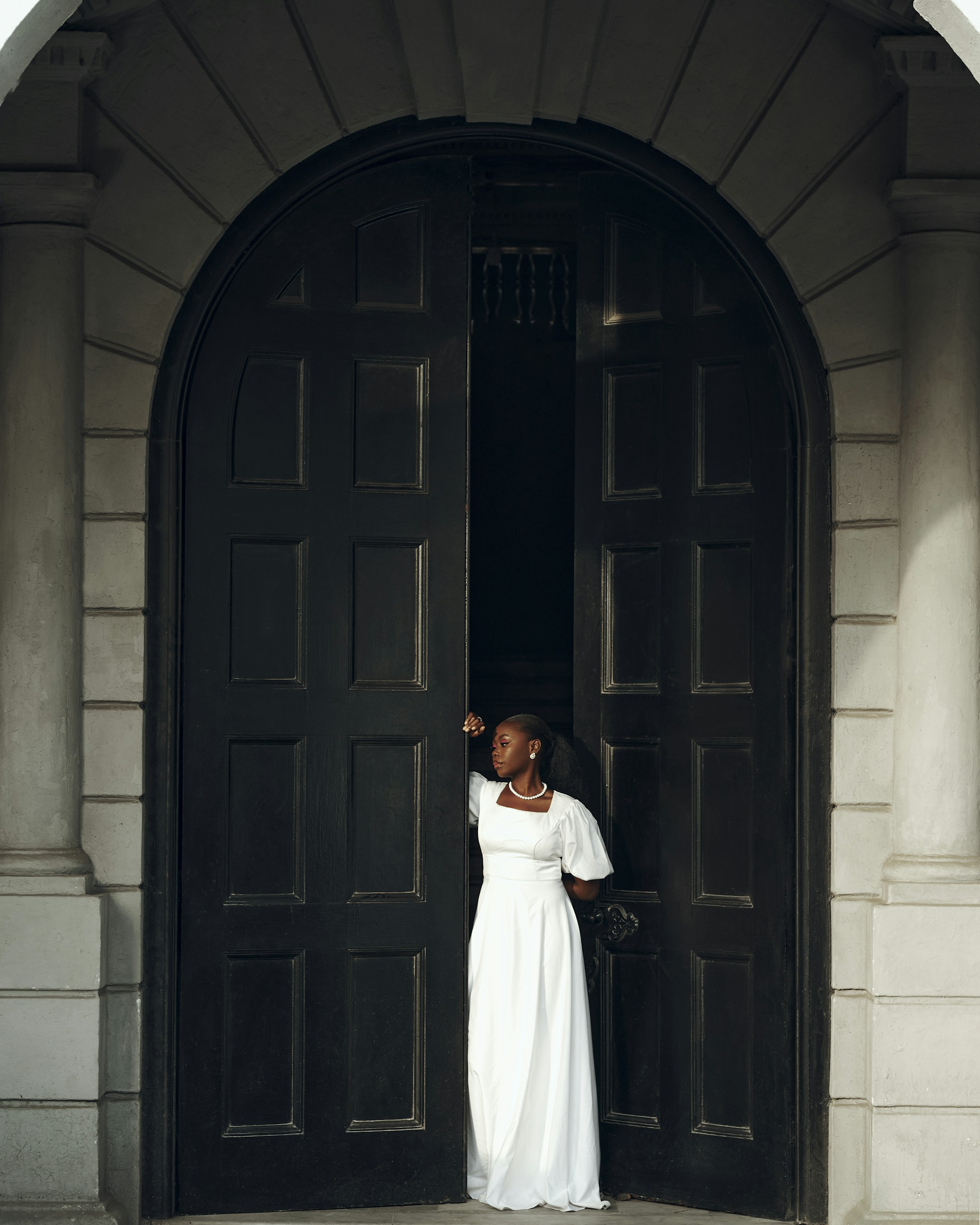 A woman in a white gown stands between tall black arched doors