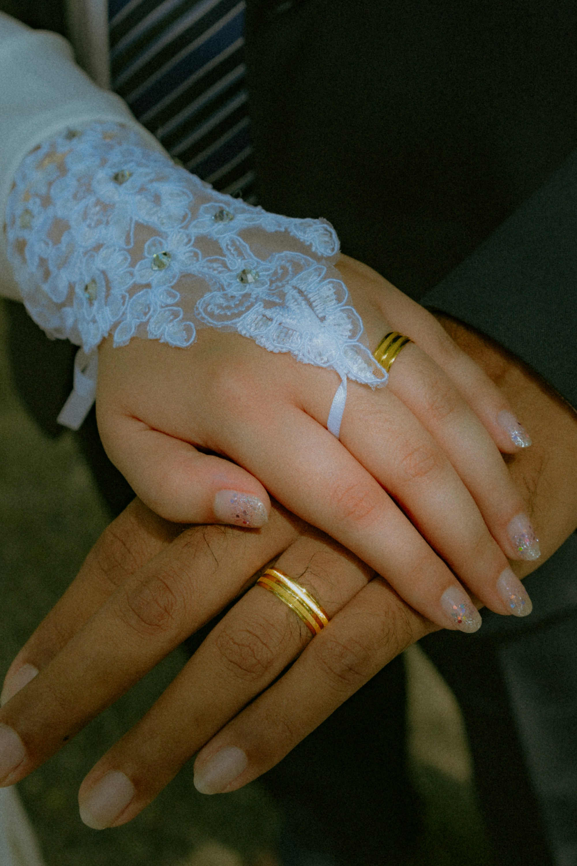 Close-up of two hands stacked together, wearing gold wedding bands; one sleeve has white lace